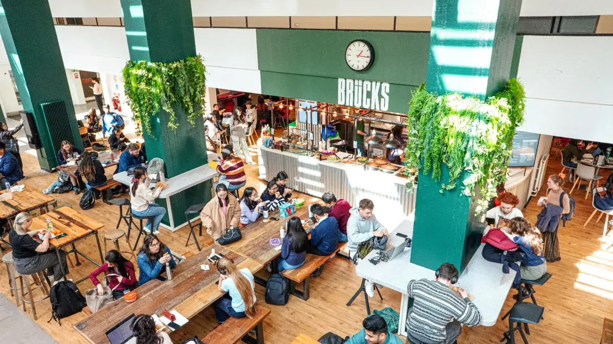 Busy interior of Brücks café at Appleton Tower with students at communal wooden tables, green columns with hanging plants and the service counter.