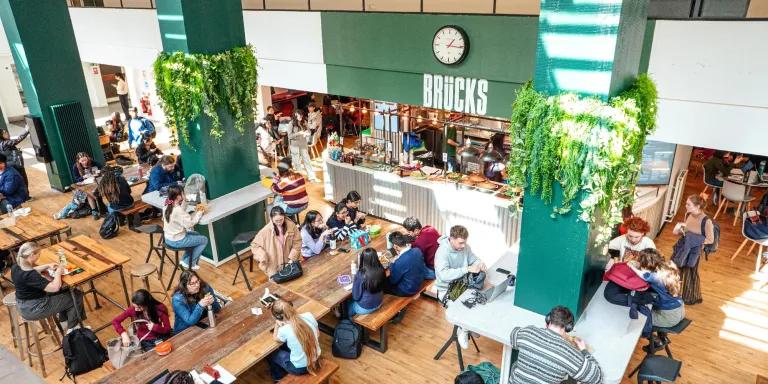 Busy interior of Brücks café at Appleton Tower with students at communal wooden tables, green columns with hanging plants and the service counter.
